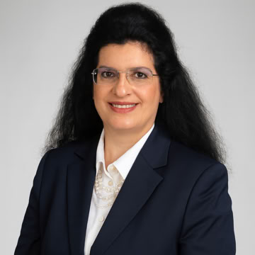 Frau mit dunklem Haar, Brille und dunklem Blazer vor grauem Hintergrund.