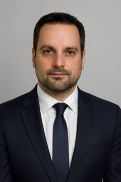 Ein Mann in einem dunklen Anzug mit weißem Hemd und dunkler Krawatte vor einem grauen Hintergrund.